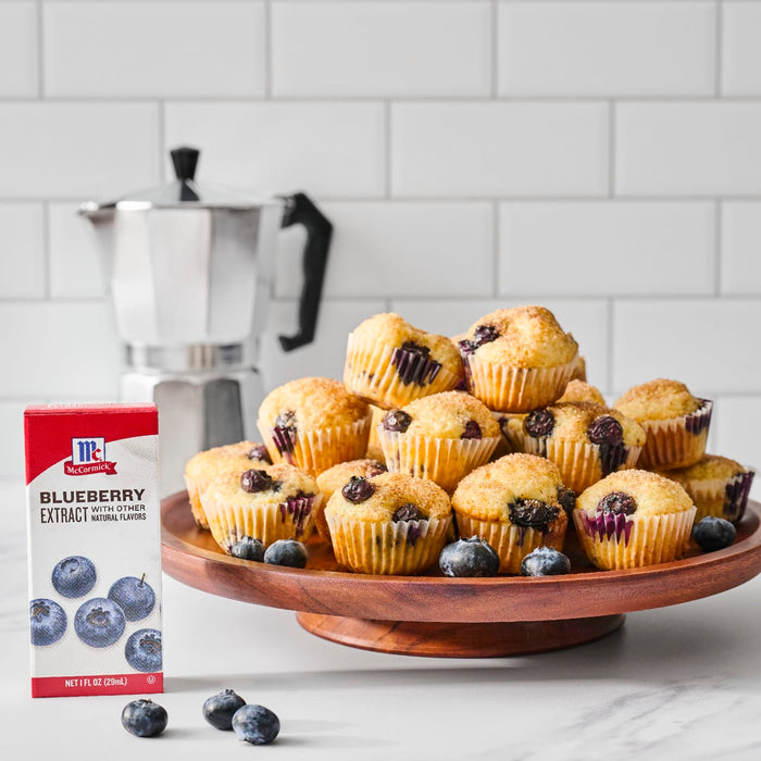A box of Mccormick Blueberry Extract 1 Fl Oz With Other Natural Flavors stands on a white counter next to a wooden cake stand overflowing with blueberry muffins, with a silver moka pot in the background.