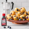 A bottle of McCormick Blueberry Extract sits next to a tower of blueberry muffins on a wooden cake stand with a Moka pot in the background.