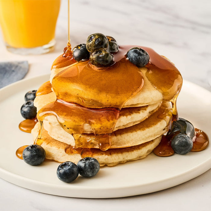A stack of pancakes topped with blueberries and syrup is being drizzled with more syrup next to a glass of orange juice.