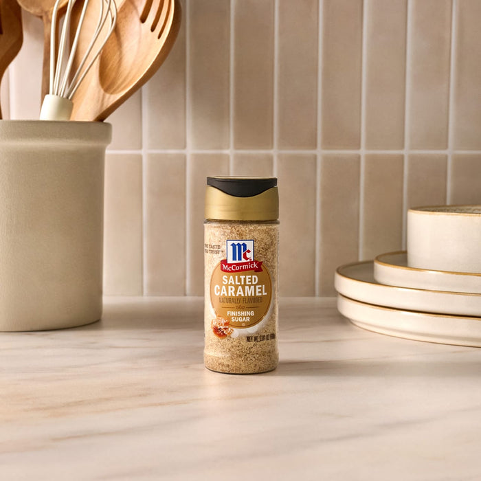 A clear glass shaker of Mccormick Salted Caramel Fin Sgr 3.81 Oz, a naturally flavored finishing sugar, is displayed on a light marble countertop with white subway tiles in the background.