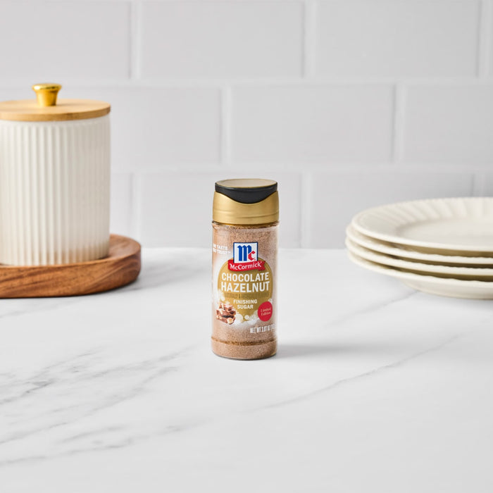 A bottle of McCormick Montreal Steak Spice stands on a marble countertop next to a stack of plates and a decorative jar.