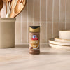 A bottle of McCormick Montreal Steak Spice stands on a marble countertop next to a stack of plates and a decorative jar.