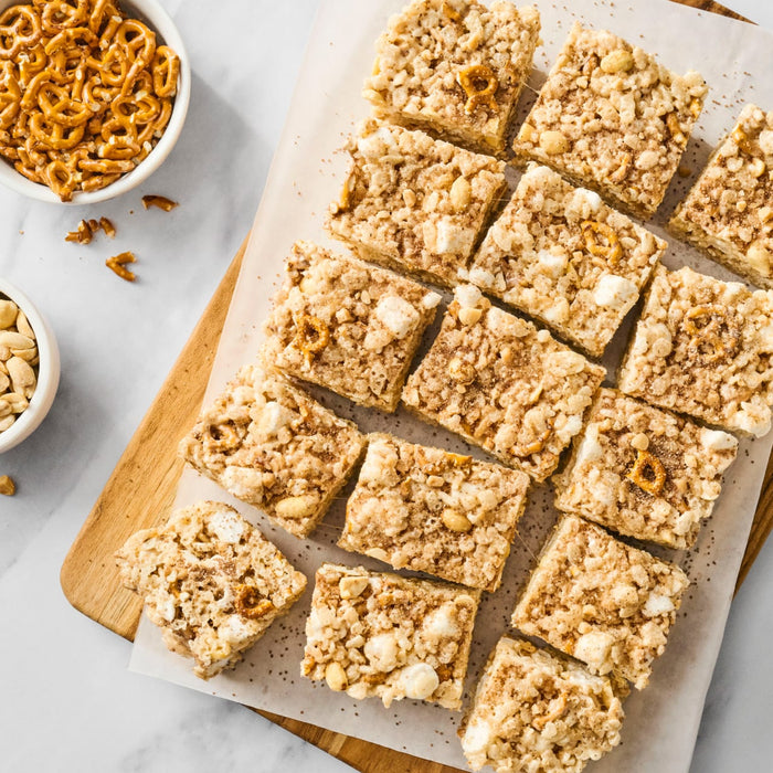 A tray of pretzel and white chocolate dessert bars sits next to bowls of pretzels and peanuts.