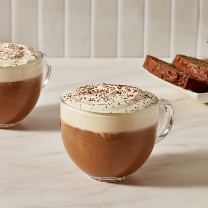 A plate of waffles topped with whipped cream and McCormick cinnamon being sprinkled on top, next to a cup of coffee.