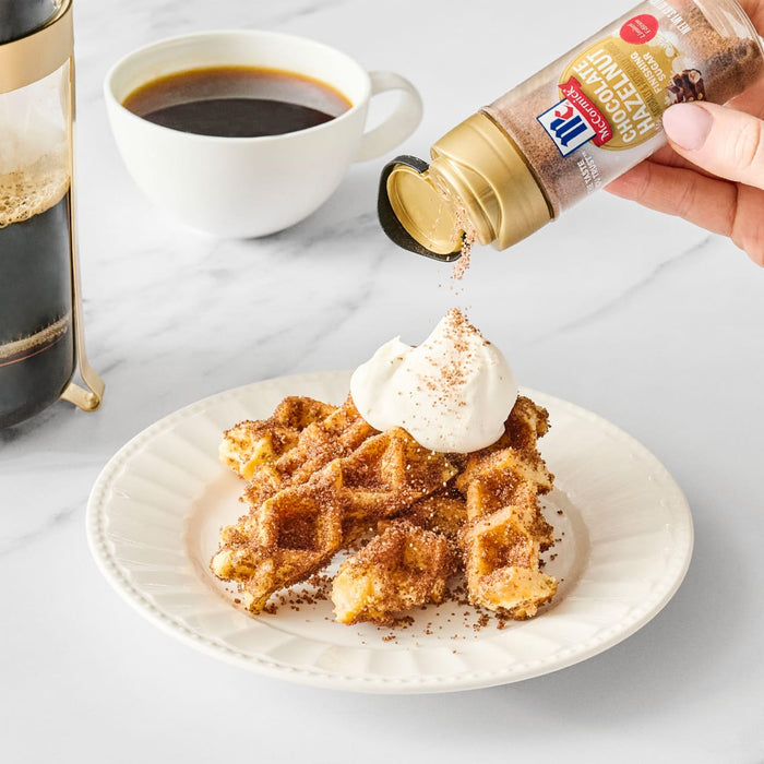 A plate of waffles topped with whipped cream and McCormick cinnamon being sprinkled on top, next to a cup of coffee.