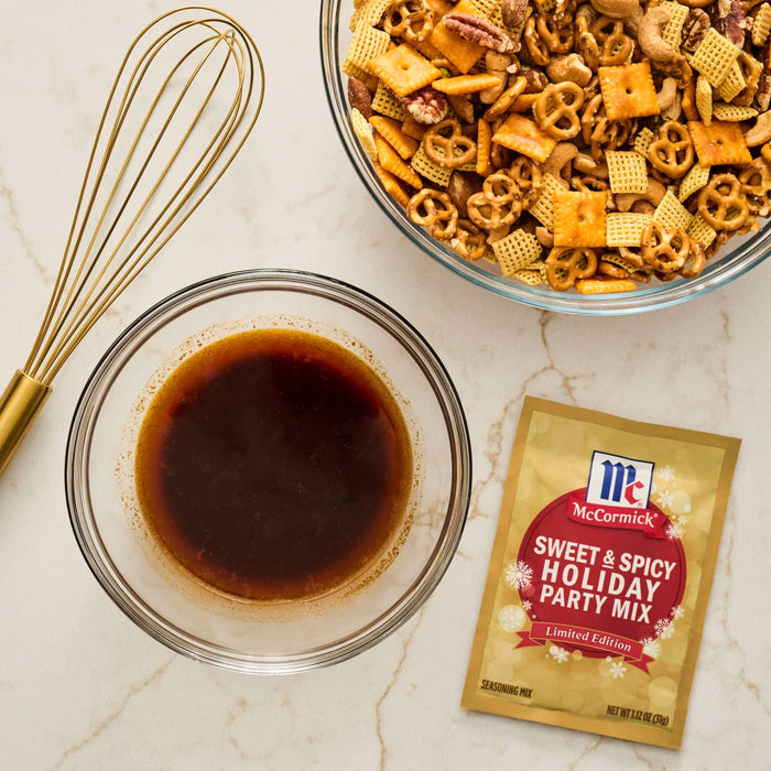 A bowl of mixed snack ingredients, a bowl of sauce, a whisk, and a packet of McCormick Sweet &amp; Spicy Holiday Party Mix are arranged on a countertop.