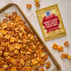 A tray of homemade party mix with pretzels, cheese crackers, and peanuts, next to a packet of McCormick Sweet &amp; Spicy Holiday Party Mix seasoning.