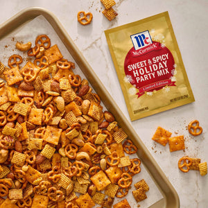 A tray of homemade party mix with pretzels, cheese crackers, and peanuts, next to a packet of McCormick Sweet &amp; Spicy Holiday Party Mix seasoning.