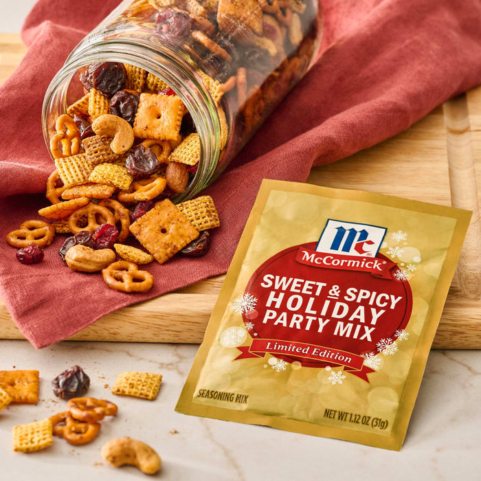 A jar of homemade snack mix spills onto a wooden board next to a packet of McCormick Sweet &amp; Spicy Holiday Party Mix seasoning.