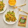Two plates of green bean casserole with McCormick Green Bean Casserole seasoning packet and a bowl of shredded cheese.