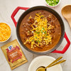 A pot of McCormick Pumpkin Chili topped with shredded cheese and green onions is displayed with a packet of McCormick Pumpkin Chili seasoning.