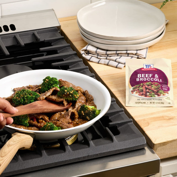 A McCormick Beef &amp; Broccoli seasoning mix packet sits next to a pan of beef and broccoli being stirred on a stovetop.