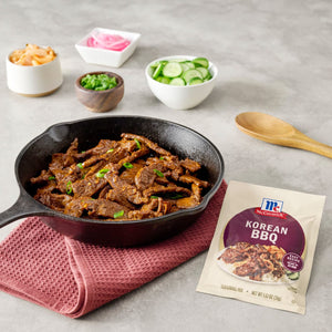 A cast iron skillet filled with Korean BBQ beef sits on a red waffle-weave towel next to a packet of McCormick Korean BBQ seasoning mix with small bowls of kimchi pickled onions green onions and cucumbers in the background.