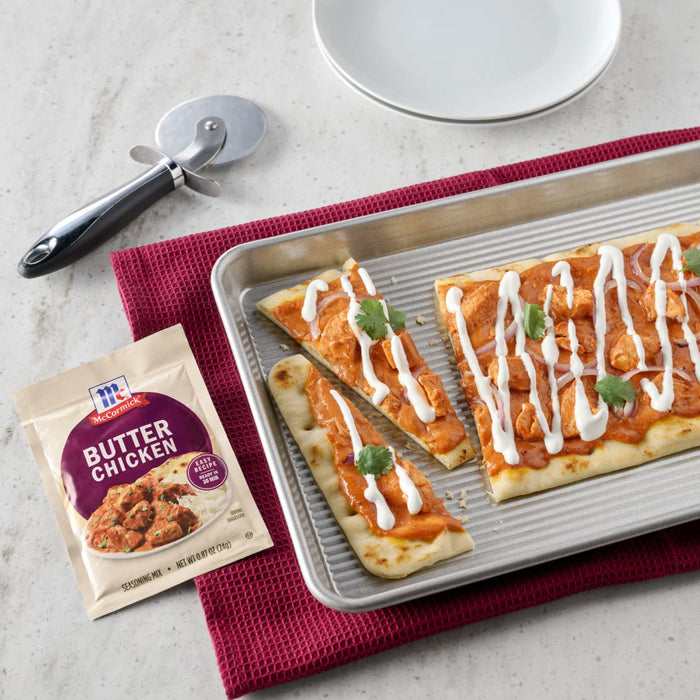 A McCormick Butter Chicken seasoning mix packet is displayed next to a baking sheet with naan bread topped with butter chicken red onions cilantro and a drizzle of white sauce.