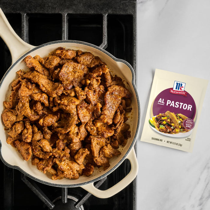 A pan of Al Pastor meat sits on a stove next to a packet of McCormick Al Pastor seasoning mix.