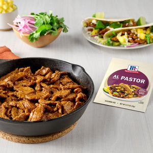 A cast iron skillet filled with Al Pastor meat accompanied by a packet of McCormick Al Pastor seasoning mix and tacos with pineapple cilantro and red onion.