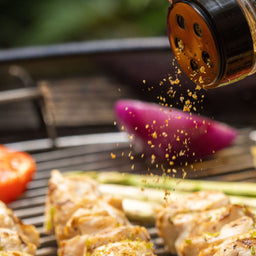 A spice shaker, specifically Mccormick Grill Mates Barbecue Seasoning, is being sprinkled over chicken skewers and vegetables on a hot grill.