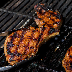A close-up view of grilled pork chops, seasoned with Mccormick Grill Mates Barbecue Seasoning, cooking on a dark barbecue grate, with one being held by metal tongs.