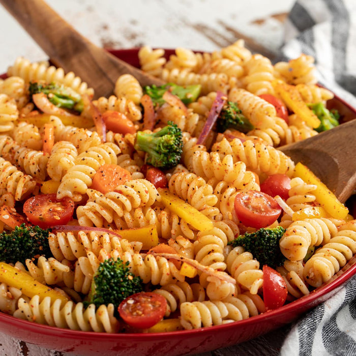 A vibrant pasta salad, seasoned with Mccormick Salad Supreme 15.75 Oz, is presented in a red bowl with wooden serving spoons, featuring fusilli pasta, broccoli, cherry tomatoes, bell peppers, and red onions