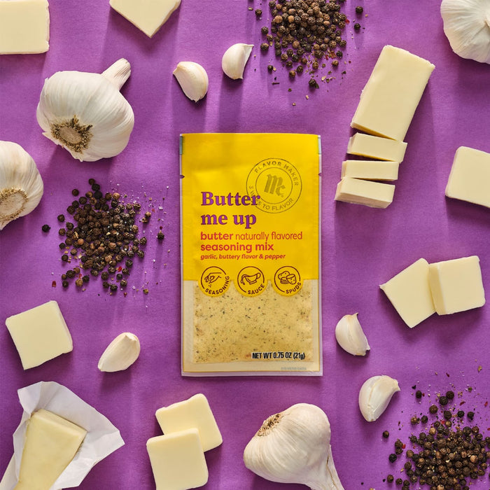 A packet of Trader Joes Butter Me Up seasoning mix is surrounded by butter garlic and peppercorns on a purple background.