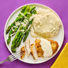 A plate of sliced breaded chicken breast with a creamy sauce mashed potatoes with gravy and asparagus drizzled with sauce showcasing a balanced and appetizing meal.