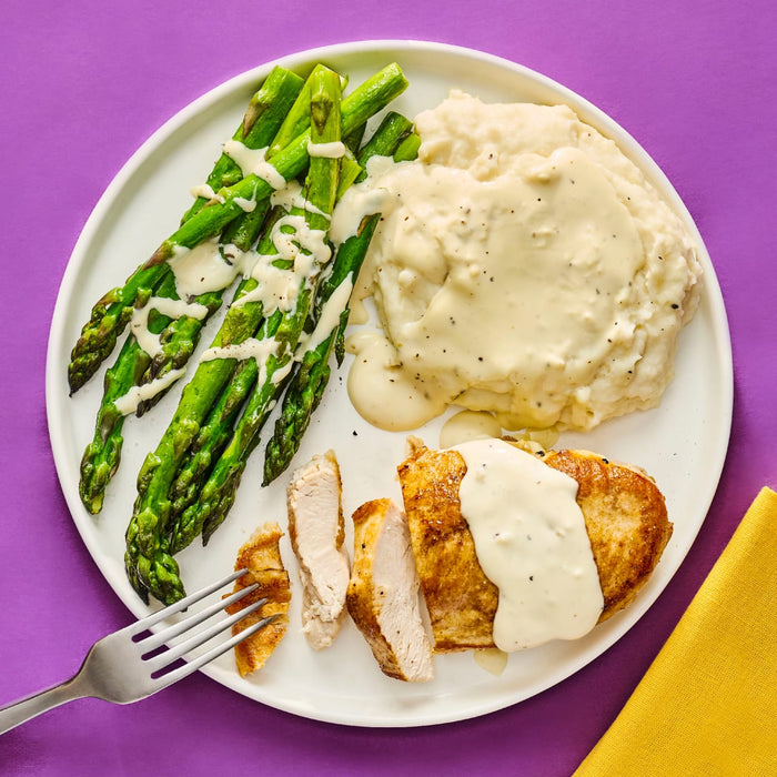 A plate of sliced breaded chicken breast with a creamy sauce mashed potatoes with gravy and asparagus drizzled with sauce showcasing a balanced and appetizing meal.