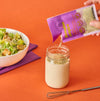 A packet of Hidden Valley Ranch seasoning is being poured into a jar of dressing with a bowl of salad and a whisk nearby.