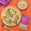 A plate of fettuccine alfredo with broccoli a bowl of shredded cheese and a packet of Flavor Maker Romaine Empire caesar seasoning mix are arranged on an orange surface.