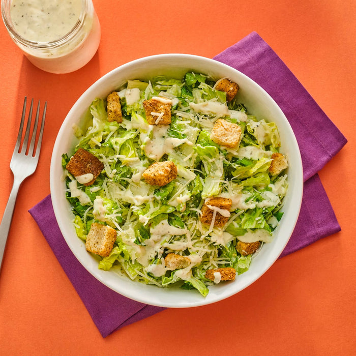 A bowl of Caesar salad with croutons and dressing set on a purple napkin against an orange background showcasing a fresh and appetizing meal.
