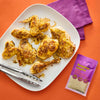 A plate of chicken seasoned with Romaine Empire Caesar seasoning mix is displayed on an orange surface next to a purple napkin and a packet of the seasoning.