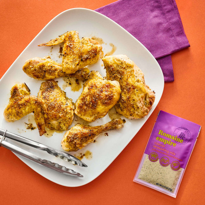 A plate of chicken seasoned with Romaine Empire Caesar seasoning mix is displayed on an orange surface next to a purple napkin and a packet of the seasoning.