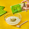 A hand pours a packet of Green Dream spice blend into a bowl of yogurt with aloe vera leaves and a wooden spoon nearby.