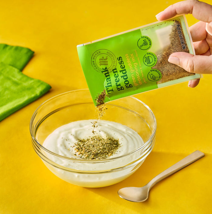 A hand pours a packet of Green Dream spice blend into a bowl of yogurt with aloe vera leaves and a wooden spoon nearby.