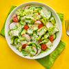 A vibrant salad featuring lettuce cucumbers radishes tomatoes and avocado dressed with a creamy dressing presented on a white plate against a bright yellow background.