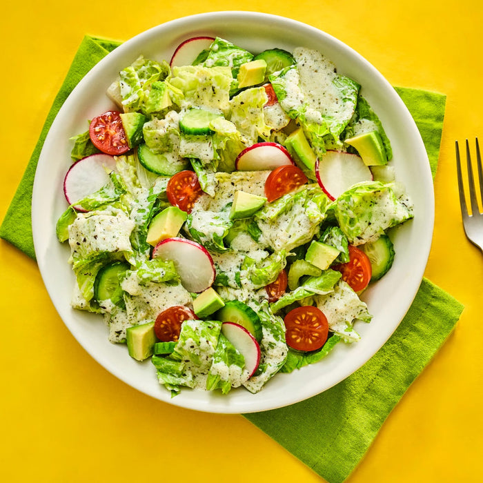 A vibrant salad featuring lettuce cucumbers radishes tomatoes and avocado dressed with a creamy dressing presented on a white plate against a bright yellow background.