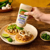 A hand squeezes a bottle of McCormick Jalapeno Mayonnaise Dressing over a shredded chicken tostada on a plate with lime wedges and a bowl of cilantro nearby.