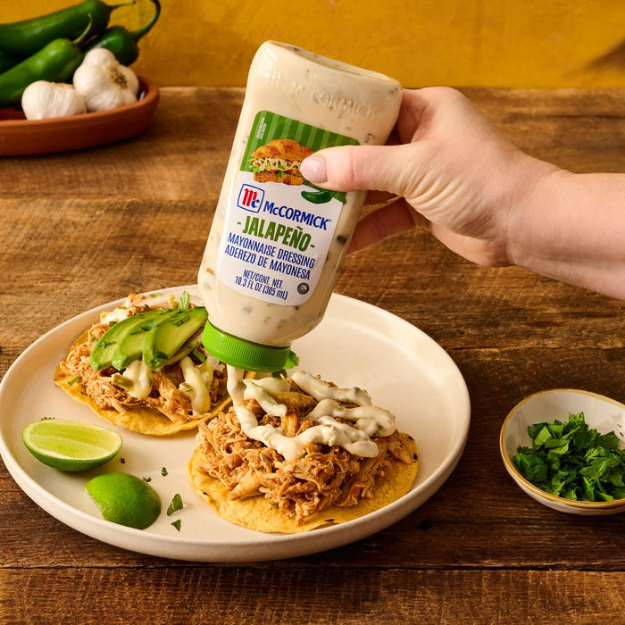 A hand squeezes a bottle of McCormick Jalapeno Mayonnaise Dressing over a shredded chicken tostada on a plate with lime wedges and a bowl of cilantro nearby.