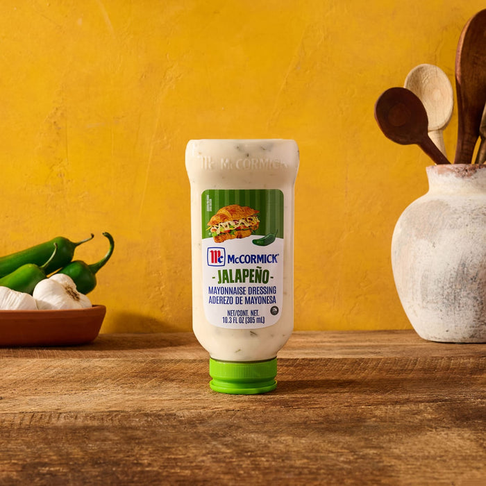 A bottle of McCormick Jalapeño Mayonnaise Dressing sits upside down on a wooden table next to fresh jalapeños and garlic against a yellow background.