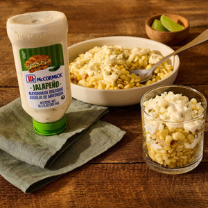 A bottle of McCormick Jalapeño Mayonnaise Dressing sits on a green napkin next to a bowl and a glass filled with corn topped with the dressing and cheese on a wooden table.