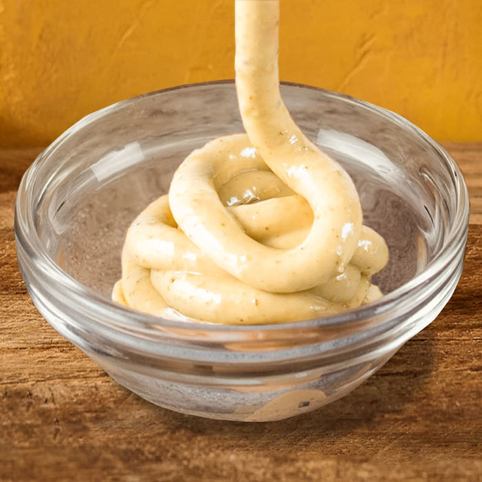 A thick creamy light brown sauce with specks of spices is being poured into a small clear glass bowl resting on a wooden table against a yellow background.
