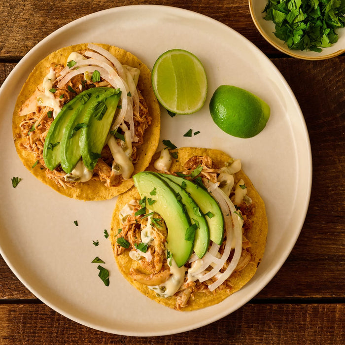 Two tacos topped with shredded meat, sliced avocado, white onions, and a creamy sauce served on a plate with lime wedges and fresh cilantro.