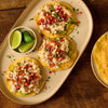 Three crispy tostadas layered with guacamole, creamy shredded chicken, and fresh salsa sit on an oval platter beside a small bowl of lime wedges on a wooden surface.