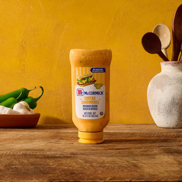 A squeeze bottle of McCormick Tortas Sandwiches Mayonnaise Dressing rests on a wooden surface beside fresh garlic and green peppers against a yellow background.