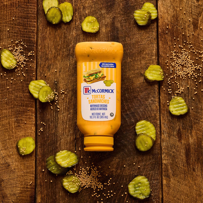 A bottle of McCormick Tortas Sandwiches Mayonnaise Dressing rests on a rustic wooden surface surrounded by scattered crinkle cut pickle slices and mustard seeds.
