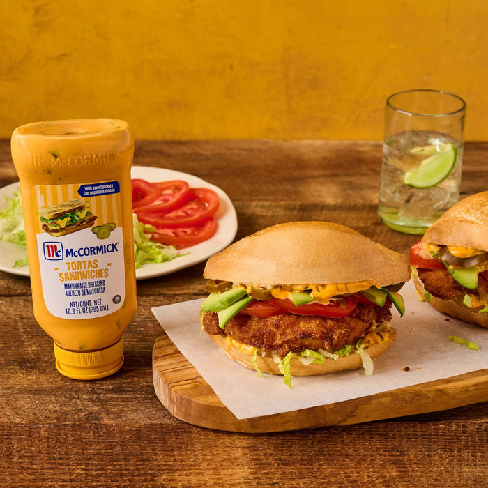 A bottle of McCormick Tortas Sandwiches Mayonnaise Dressing sits on a rustic wooden table next to a hearty torta sandwich layered with breaded meat, avocado, tomato, and the creamy dressing.