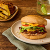 A Mexican style torta sandwich layered with seasoned meat, grilled pineapple, avocado, and melted cheese rests on a speckled plate upon a rustic wooden table.