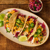 Four appetizing fish tacos topped with vibrant purple cabbage slaw, jalapeno slices, cilantro, and a creamy orange sauce are arranged on an oval plate with lime wedges alongside a small bowl of extra slaw on a rustic wooden table.