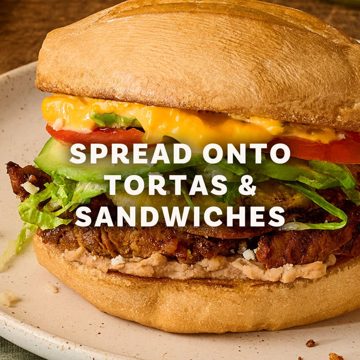 A close up of a torta filled with meat, beans, lettuce, tomato, and a yellow sauce with overlaid text reading SPREAD ONTO TORTAS AND SANDWICHES.
