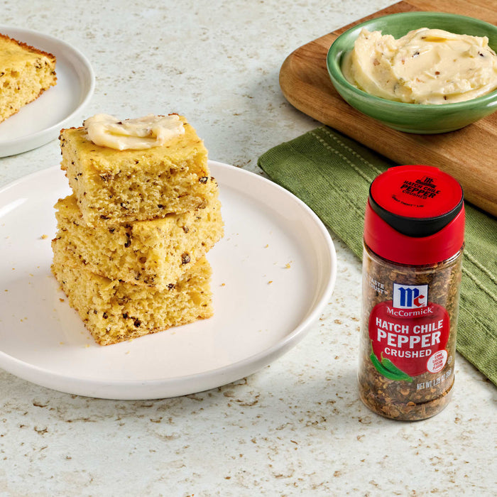 A stack of hatch chile pepper cornbread squares topped with butter sits on a plate next to a McCormick Hatch Chile Pepper Crushed spice container.