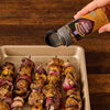 A hand sprinkles Mccormick Grill Mates Bourbon &amp; Oak Seasoning Ground 3.37 Oz over freshly grilled meat and vegetable skewers on a baking sheet.